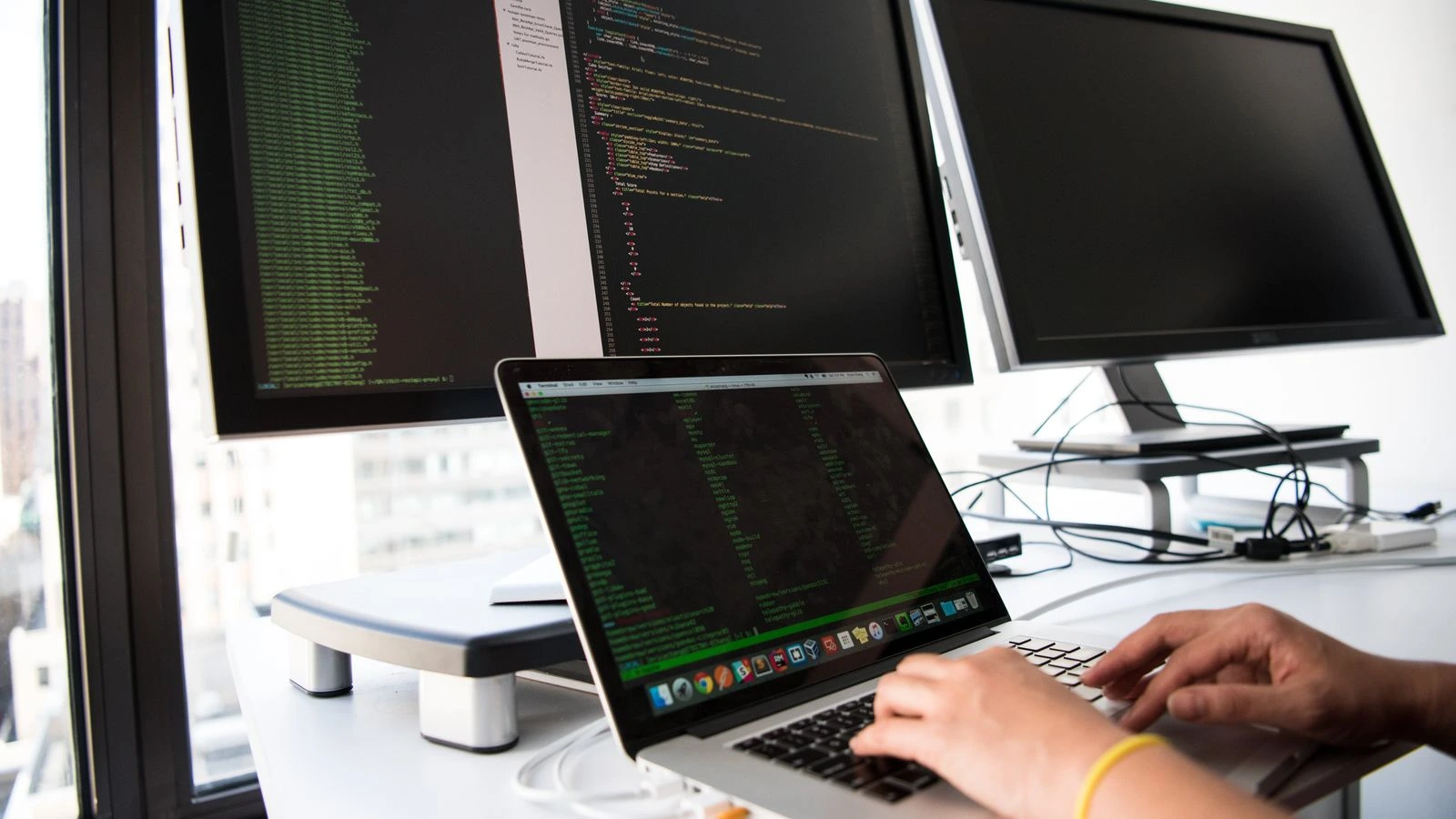 A developer writes code on a laptop in front of multiple monitors in an office setting